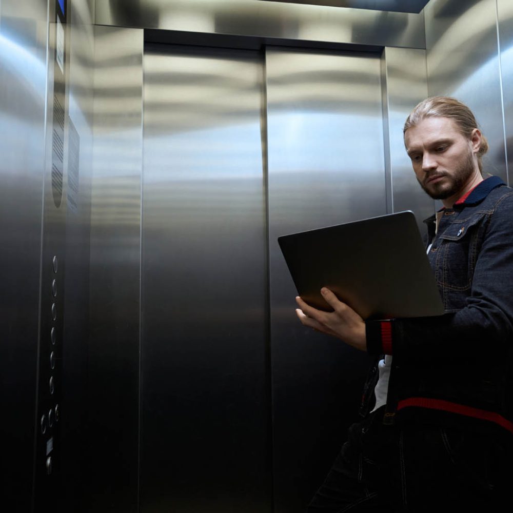 Young man is in the cabin of a passenger elevator, he is chatting online on a laptop